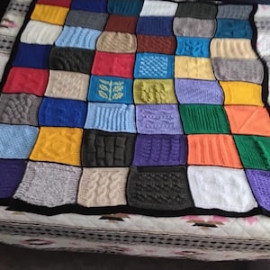 May include: A multi-colored patchwork blanket composed of numerous square knitted panels. Each square features a unique textured pattern, including cables, ribs, and geometric designs in shades of red, blue, green, yellow, purple, orange, and white.