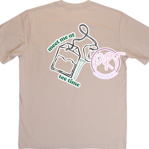 Könnte beinhalten: Beigefarbenes T-Shirt mit einer Grafik eines Teebeutels und dem grünen Schriftzug "meet me at tee time". Das Design enthält ein kreisförmiges Logo. Das Shirt besteht aus einem weichen Material.
