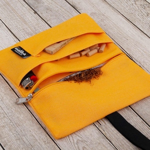May include: A yellow canvas tobacco pouch with a zipper closure and a black strap. The pouch has three compartments, one for tobacco, one for filters, and one for rolling papers. The pouch also has a small pocket for a lighter. The pouch is made by "tabak-tasche" and is handmade.