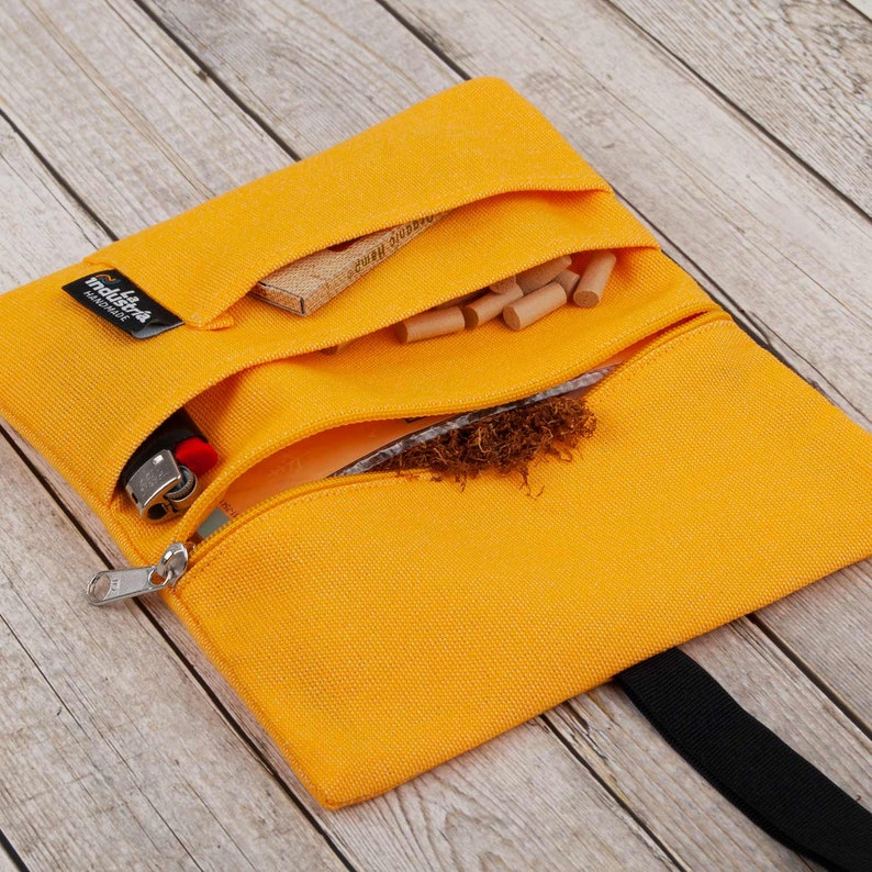May include: A yellow canvas tobacco pouch with a zipper closure and a black strap. The pouch has three compartments, one for tobacco, one for filters, and one for rolling papers. The pouch also has a small pocket for a lighter. The pouch is made by "tabak-tasche" and is handmade.