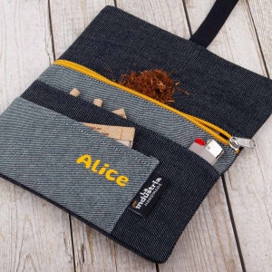 Könnte beinhalten: Eine schwarze Tabaktasche aus Denim mit einem gelben Reißverschluss und einem gelben Etikett mit der Aufschrift "la industria handmade". Die Tasche hat ein Fach für ein Feuerzeug und ein Fach für Drehpapier. Die Tasche ist mit dem Namen "Alice" in gelbem Garn personalisiert.