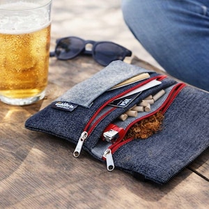 Può includere: Un astuccio in denim con cerniere rosse, contenente tabacco, sigarette e un accendino. Un bicchiere di birra e occhiali da sole sono sullo sfondo. L'astuccio ha una patta in feltro grigio e un'etichetta "ackistrie handmade".