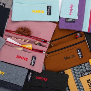 May include: A collection of personalised zippered pouches in various colours and fabrics. The pouches feature embroidered names such as "Daniel", "Maria", "Sylvia", "Paul", "Anne", "Moritz", "Stefan", "Alice", "Alex", and "Lucie".