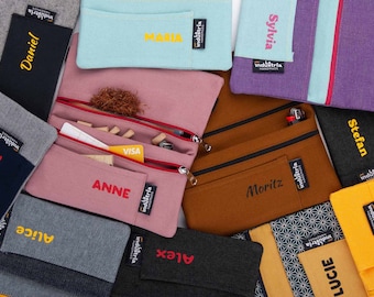 Puede incluir: Una colección de estuches con cremallera personalizados en varios colores y tejidos. Los estuches presentan nombres bordados como "Daniel", "Maria", "Sylvia", "Paul", "Anne", "Moritz", "Stefan", "Alice", "Alex" y "Lucie".