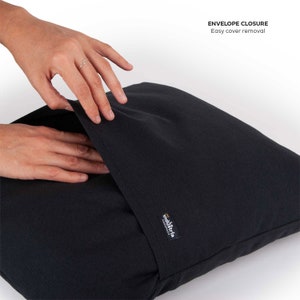 Puede incluir: Una almohada negra con cierre de sobre. La almohada tiene una etiqueta que dice "industria design". La almohada est&aacute; siendo sostenida por una mano, mostrando lo f&aacute;cil que es quitar la funda.