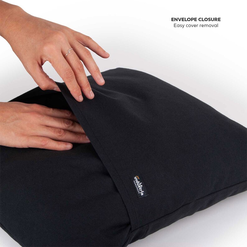 Puede incluir: Una almohada negra con cierre de sobre. La almohada tiene una etiqueta que dice "industria design". La almohada est&aacute; siendo sostenida por una mano, mostrando lo f&aacute;cil que es quitar la funda.