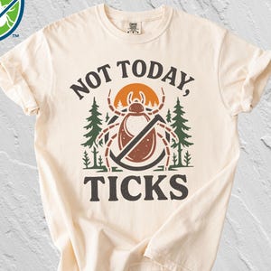 May include: Cream-colored t-shirt with the words "NOT TODAY, TICKS" in a bold, dark brown font. The design includes a tick illustration with a line through it, a rising sun, and pine trees. A shield logo is in the upper left corner.