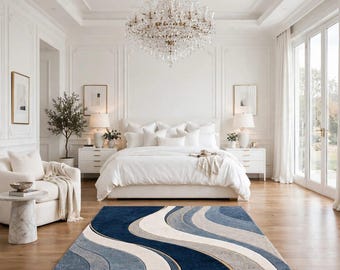May include: A rectangular area rug featuring a wavy, abstract pattern in shades of navy blue, light blue, gray, and cream. The rug is positioned on a light-colored hardwood floor in a bedroom with a white bed, white nightstands, and a chandelier.