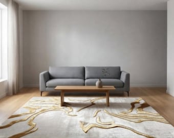 May include: A rectangular area rug featuring a light gray base with raised, flowing gold-colored abstract lines that create a textured, organic pattern across the surface. The rug is positioned on a wooden floor in front of a gray sofa and table.