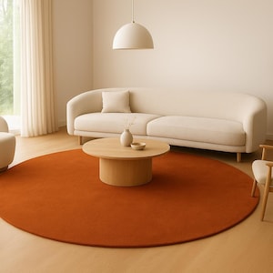 Puede incluir: Un salón moderno con una gran alfombra redonda de color óxido. La habitación cuenta con un sofá color crema, un sillón y una mesa de centro de madera. Una pequeña mesa auxiliar con una planta y una lámpara colgante completan la escena.