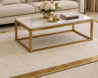 May include: A rectangular area rug featuring a cream-colored center and a wide, textured gold border. The rug is positioned on a light-colored floor, providing a neutral base that complements the surrounding furniture and the gold-framed coffee table.