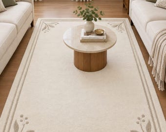 May include: A rectangular area rug in a cream color featuring a double-line border and a botanical leaf motif in each corner. The rug is made of a soft, textured material and is positioned on a wooden floor between two light-colored sofas.