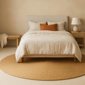Puede incluir: Un dormitorio con una cama con cabecero beige, almohadas y funda n&oacute;rdica. Un coj&iacute;n naranja a&ntilde;ade un toque de color. Una alfombra redonda y tejida en un tono c&aacute;lido est&aacute; en el suelo. Una mesita de noche de madera con una l&aacute;mpara est&aacute; a la derecha.