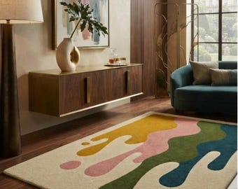 May include: A rectangular area rug featuring a cream-colored base with large, fluid, abstract shapes in mustard yellow, soft pink, forest green, and deep teal. The design mimics dripping paint with rounded edges and scattered circular spots.