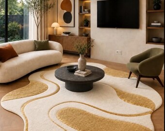 May include: An irregular, organic-shaped area rug featuring a cream-colored base with flowing, wavy patterns in a mustard yellow hue. The rug has a thick, textured pile and is positioned on a light-colored wooden floor in a living room setting.