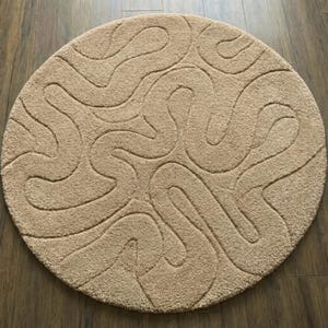 Puede incluir: Una alfombra redonda beige con un patrón ondulado en relieve. El diseño presenta líneas curvas continuas que crean una superficie texturizada. La alfombra está colocada sobre un suelo de madera, proporcionando una estética neutra y cálida. Diámetro: aprox. 150 cm.
