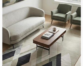 May include: A rectangular area rug featuring a geometric pattern of triangles in shades of charcoal, sage green, and cream. The rug is positioned on a light wood floor beneath a wooden coffee table, a light gray curved sofa, and two sage green armchairs.