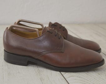 Puede incluir: Un par de zapatos derby de cuero marrón con acabado liso, punteras redondeadas, cordones de color marrón oscuro y un tacón bajo. Cada zapato contiene una horma de plástico beige para mantener la forma de la estructura del calzado.
