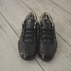 May include: Pair of black Y-3 sneakers. The shoes have a leather upper with a glossy finish. The laces are black, and the tongue features the Y-3 logo. The shoes are set against a light-colored wooden surface.