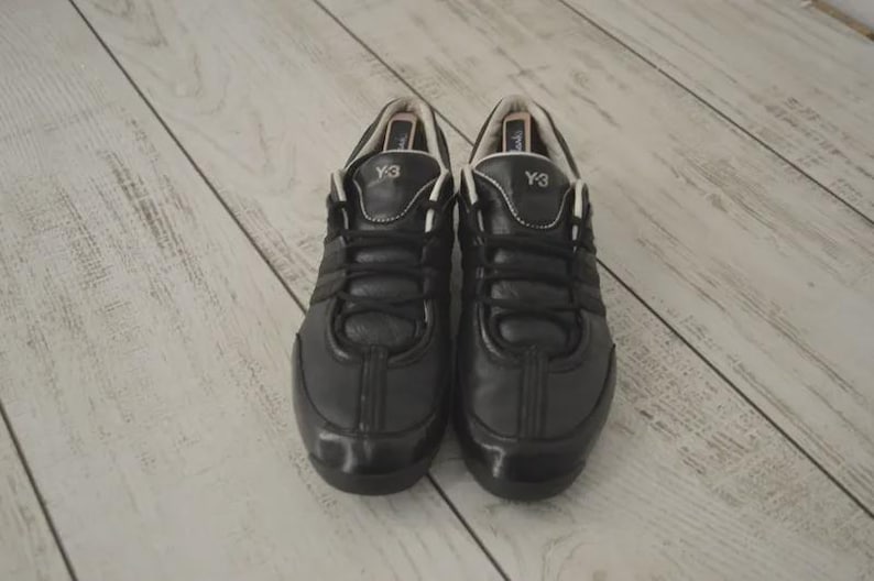 May include: Pair of black Y-3 sneakers. The shoes have a leather upper with a glossy finish. The laces are black, and the tongue features the Y-3 logo. The shoes are set against a light-colored wooden surface.