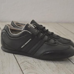 May include: Black leather sneakers with the text "YOHJI YAMAMOTO" on the side. The shoes have a low profile design with a textured toe cap and three stripes. The laces are black.