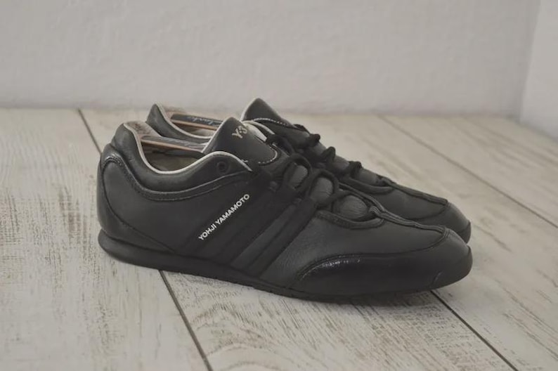 May include: Black leather sneakers with the text "YOHJI YAMAMOTO" on the side. The shoes have a low profile design with a textured toe cap and three stripes. The laces are black.