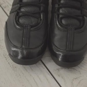 May include: Close-up of two black leather sneakers. The shoes have a glossy finish on the toe and a matte finish on the rest of the shoe. The laces are black, and the shoes are on a light-colored wooden surface.