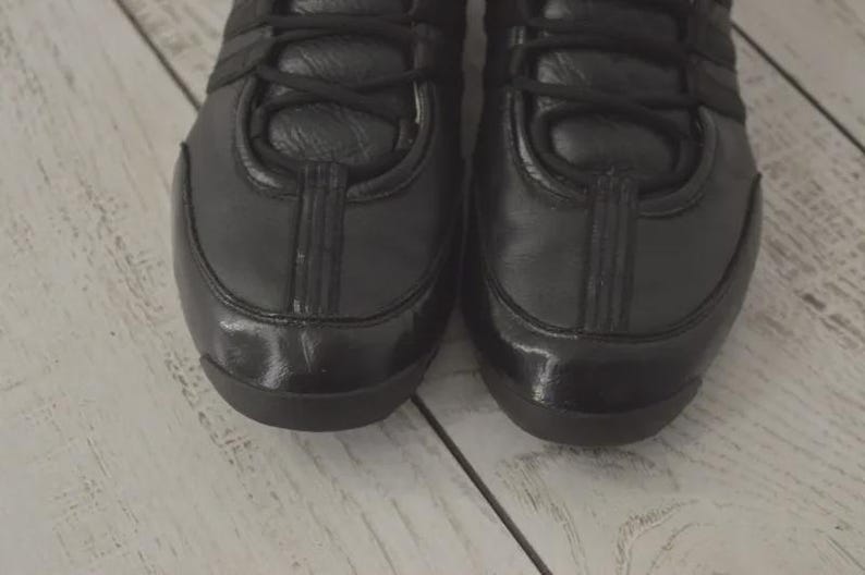 May include: Close-up of two black leather sneakers. The shoes have a glossy finish on the toe and a matte finish on the rest of the shoe. The laces are black, and the shoes are on a light-colored wooden surface.