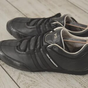 May include: Pair of black leather sneakers with the Y-3 logo and three stripes. The shoes have a low profile design with a rounded toe and a lace-up closure. The brand name "YOHJI YAMAMOTO" is visible on the side.