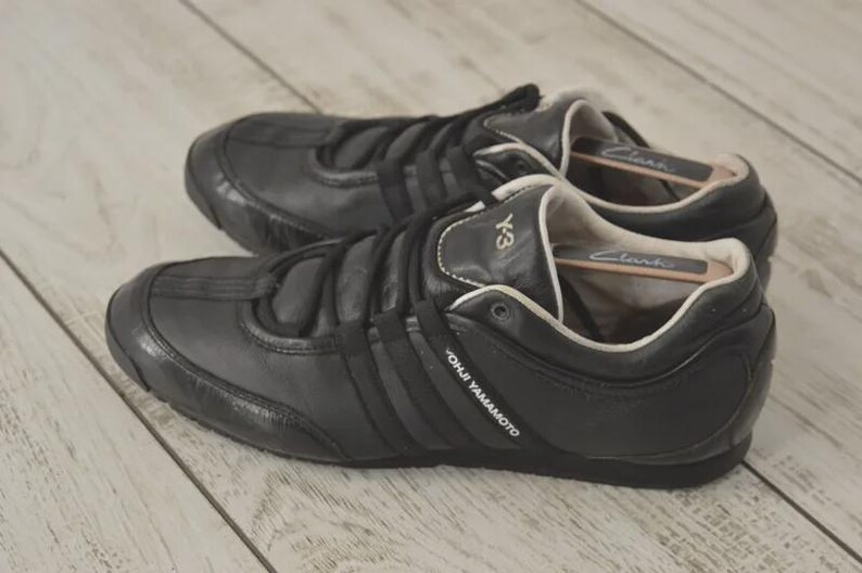 May include: Pair of black leather sneakers with the Y-3 logo and three stripes. The shoes have a low profile design with a rounded toe and a lace-up closure. The brand name "YOHJI YAMAMOTO" is visible on the side.