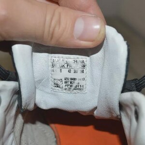 May include: Close-up of the inside of a black and white Adidas sneaker. The shoe's tongue features a label with size and manufacturing information. The interior is white with an orange sole.