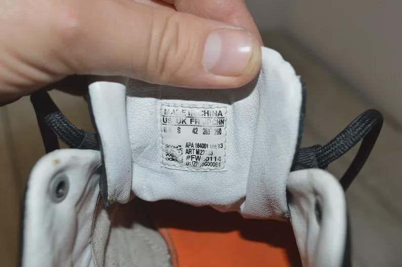 May include: Close-up of the inside of a black and white Adidas sneaker. The shoe's tongue features a label with size and manufacturing information. The interior is white with an orange sole.