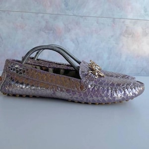 Peut inclure: Un mocassin violet métallique avec un motif texturé rappelant la peau de serpent, doté d'une garniture en métal doré sur le dessus et d'une semelle à picots en caoutchouc. La chaussure contient un embauchoir argenté pour le maintien.