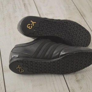 May include: Black leather sneakers with a low profile. The shoes feature three stripes on the side and the Y-3 logo in gold on the sole. The soles have a geometric pattern.