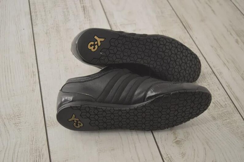 May include: Black leather sneakers with a low profile. The shoes feature three stripes on the side and the Y-3 logo in gold on the sole. The soles have a geometric pattern.