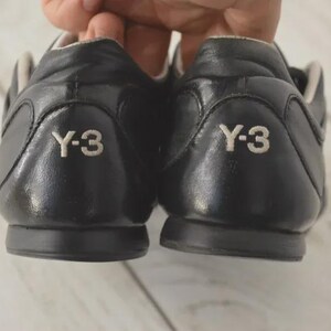 May include: A pair of black leather sneakers with the white "Y-3" logo on the heel. The shoes have a low-profile design with a rounded toe and a contrasting light-colored interior.
