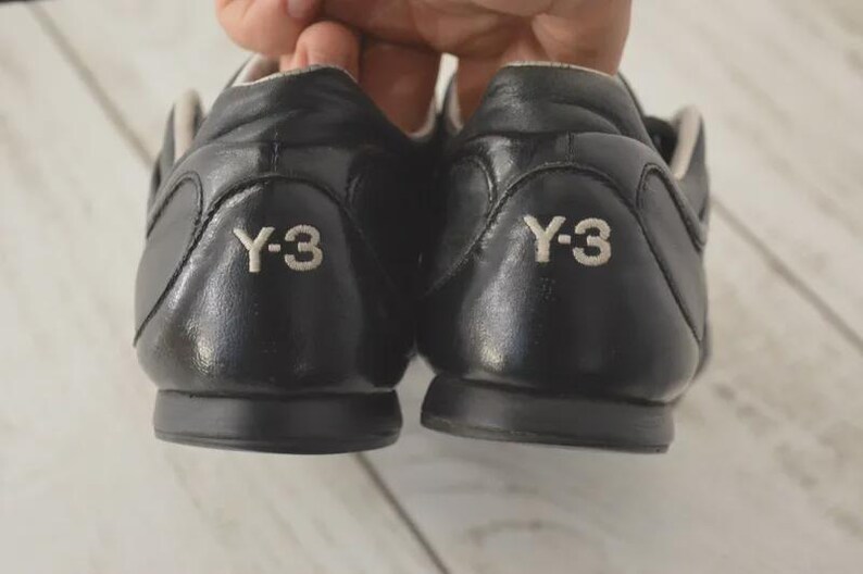 May include: A pair of black leather sneakers with the white "Y-3" logo on the heel. The shoes have a low-profile design with a rounded toe and a contrasting light-colored interior.