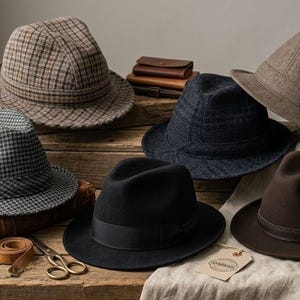 Puede incluir: Una colección de seis sombreros fedora en varios colores y patrones, incluyendo cuadros marrones, pata de gallo gris, negro sólido, azul oscuro y marrón sólido. Los sombreros están dispuestos sobre una superficie de madera junto a una cinta métrica y tijeras.
