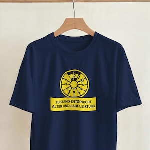 Puede incluir: Camiseta azul marino con un diseño gráfico amarillo en forma de reloj y el texto "ZUSTAND ENTSPRICHT ALTER UND LAUFLEISTUNG". La camiseta está colgada en una percha. El diseño está centrado en la parte delantera.