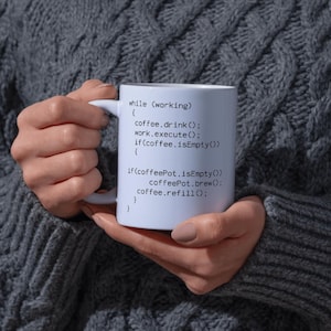 Peut inclure: Mug en céramique blanche avec une anse, affichant du code de programmation en texte noir. Le code comprend des commandes telles que "coffee.drink()" et "coffee.refill()". Le mug est tenu par une personne portant un pull gris.