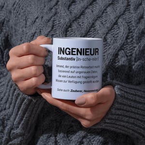 Peut inclure: Mug en céramique blanche avec le mot "INGENIEUR" en noir et une définition en allemand. La tasse est tenue par une personne portant un pull gris. La tasse a une anse blanche et est remplie d'un liquide inconnu.