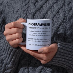 Könnte beinhalten: Eine weiße Keramik-Tasse mit dem Wort "PROGRAMMIERER" in Schwarz aufgedruckt, zusammen mit einer Definition auf Deutsch. Die Tasse wird von einer Person gehalten, die einen grauen Pullover trägt.