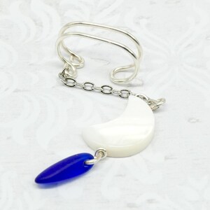 May include: A silver-toned ear cuff with a chain and a white crescent moon charm, accented by a blue teardrop-shaped bead. The jewelry piece is set against a white patterned background.