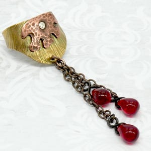 May include: A handcrafted ring with a mixed metal design. The ring features a textured brass band, a copper accent with a small crystal, and a chain with three red glass teardrop charms. A unique piece of jewelry.