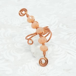 May include: A copper-colored wire ring with peach-colored beads. The ring features a unique design with a spiral at the top and bottom, and the beads are strung along the side. The ring is set against a white background with a subtle floral pattern.