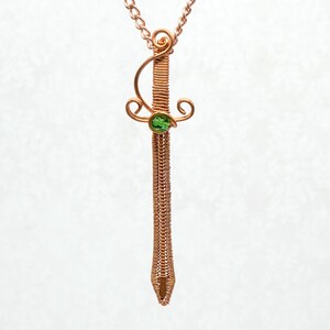 May include: Copper wire pendant necklace in the shape of a sword. The sword features a green gemstone and decorative swirls. The pendant hangs from a copper chain against a white background.