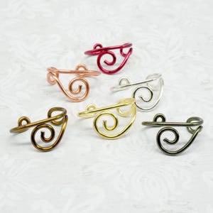 May include: A collection of six wire rings in various metallic colors. The rings feature a swirling, open design. Colors include red, copper, silver, gold, bronze, and dark gray. The rings are displayed on a white patterned surface.