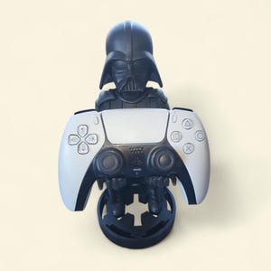 May include: A black Darth Vader-themed PlayStation 5 controller stand. The stand features a detailed Darth Vader bust holding a white and black PS5 controller. The base is black with a cross-shaped design, providing a secure spot for the controller.
