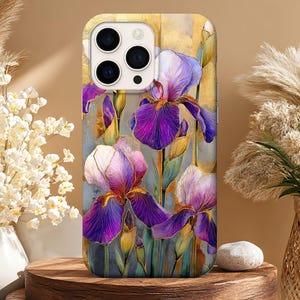 May include: A phone case featuring a watercolor painting of purple and white iris flowers. The case has a gold and gray background. The phone case is displayed on a wooden stand with decorative flowers in the background.