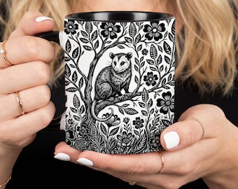 May include: A black and white ceramic mug featuring a detailed illustration of an opossum perched on a tree branch, surrounded by a dense pattern of stylized flowers and leaves. The mug has a solid black interior and a matching black handle.